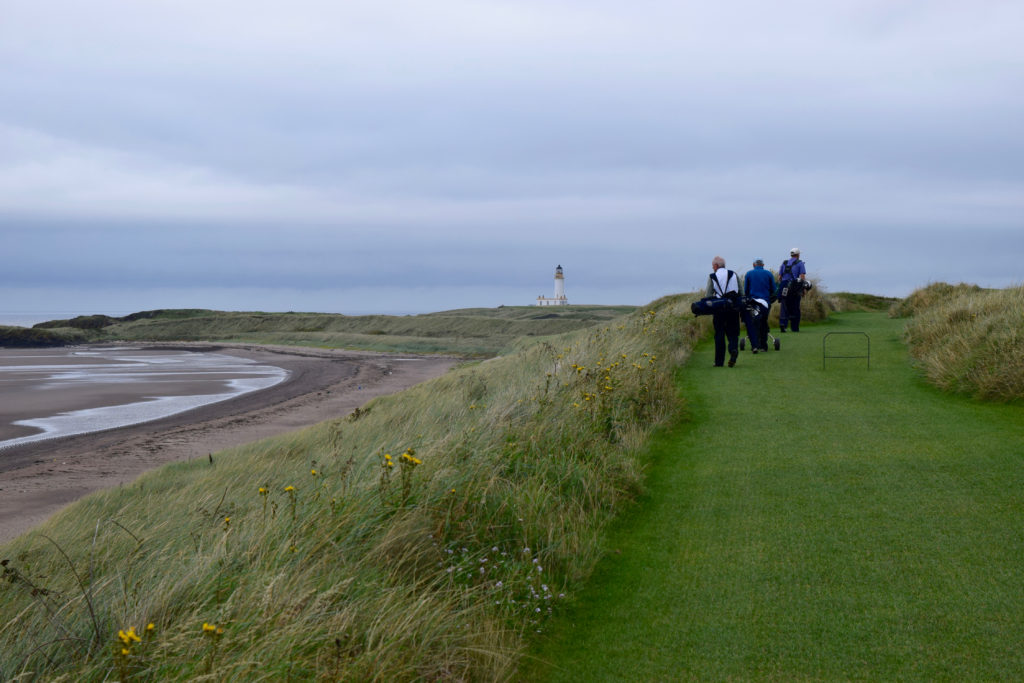Tom Cade, Blaine Newnham and Rob Perry walk from the fifth green to the sixth tee at Trump Turnberry Golf Club, with Turnberry Lighthouse in the distance.